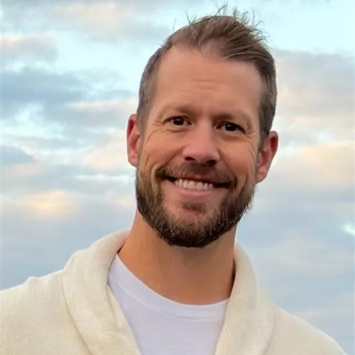 A man wearing a white sweater and a white t-shirt is standing outdoors against a backdrop of a cloudy sky during daylight. The scene captures a serene and natural atmosphere with soft lighting. The focus is on the upper body and clothing details.